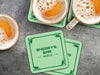 Custom square pulpboard coasters are shown sitting on a dark gray stone background with beer mugs surrounding them. The beer mugs have coasters underneath them. The custom coasters feature a green golf bar design with interlocking golf clubs used as a border. The bar name and location can be customized.
