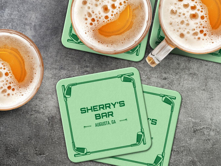 Custom square pulpboard coasters are shown sitting on a dark gray stone background with beer mugs surrounding them. The beer mugs have coasters underneath them. The custom coasters feature a green golf bar design with interlocking golf clubs used as a border. The bar name and location can be customized.
