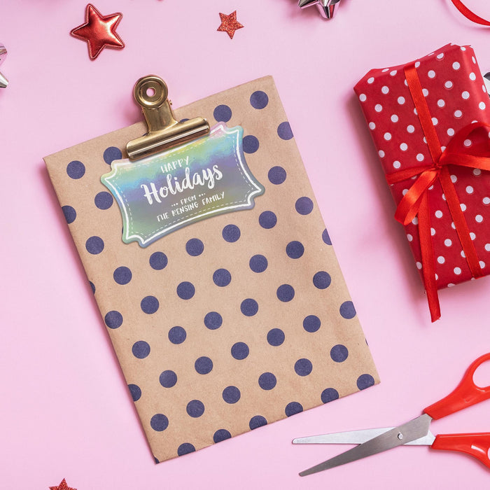 Personalized Happy Holidays slim rectangle gift tag clipped to a polka dot kraft gift bag on a pink holiday background with wrapped presents and scissors.