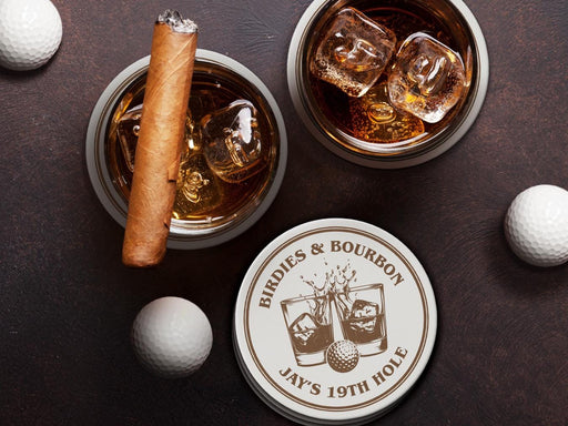 Two whiskey glasses are shown sitting on custom coasters on a dark brown table while surrounded by three white golf balls. An unlit cigar sits on top of one of the glasses. A stack of custom coasters can also be seen near the glasses. The custom coasters feature a golf ball centered between two whiskey glasses clinking together and splashing out with customizable text wrapping around the illustration. The coaster design is printed in brown.
