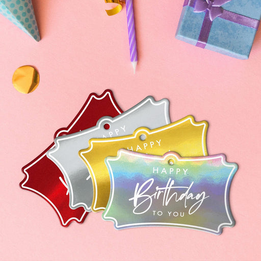 Four birthday tags in metallic gold, metallic silver, metallic red, and metallic rainbow lying beside colorful gifts and party decorations.
