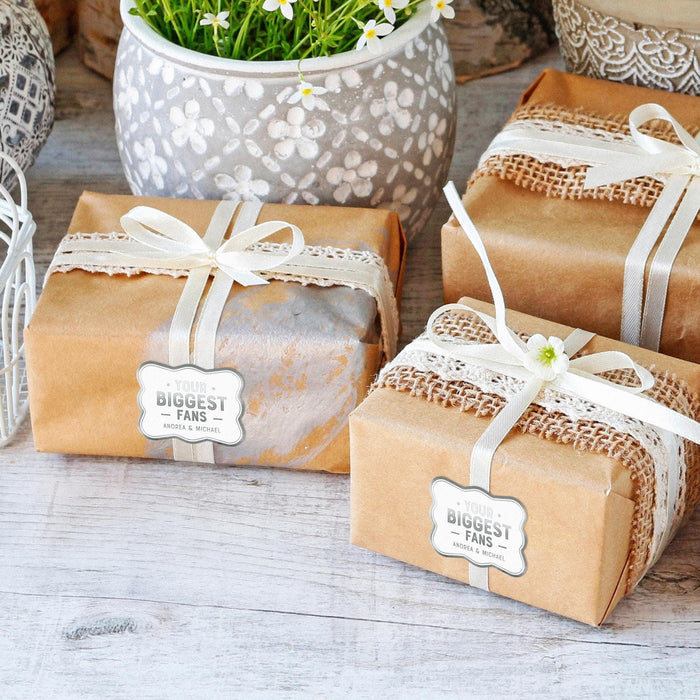 Small wrapped gift boxes decorated with personalized “Your Biggest Fans” wedding gift tags in white foil. Styled as elegant wedding favors and bridal shower gift packaging.