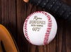 An engraved baseball sits on a dark wooden table besides a baseball bat and baseball glove. The baseball has a player's name, team name, and number engraved into it. This design features stylized text using script and 3D block lettering.