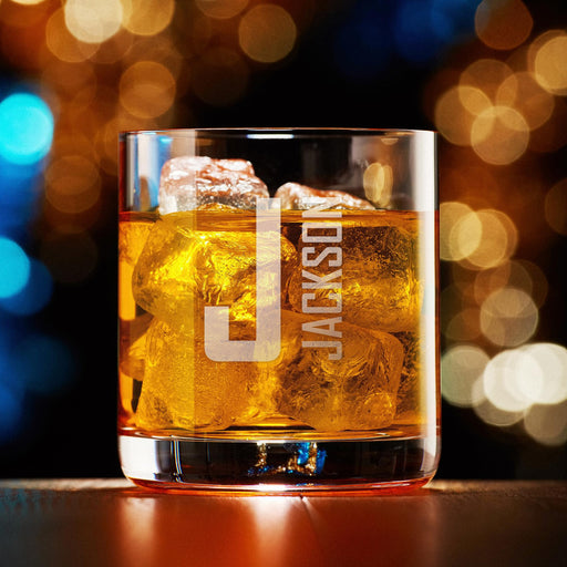Elegant product photo of a personalized whiskey glass with a large “J” initial and vertical “Jackson” name engraving, set against a warm bokeh background. The glowing lights highlight the modern and bold custom design.