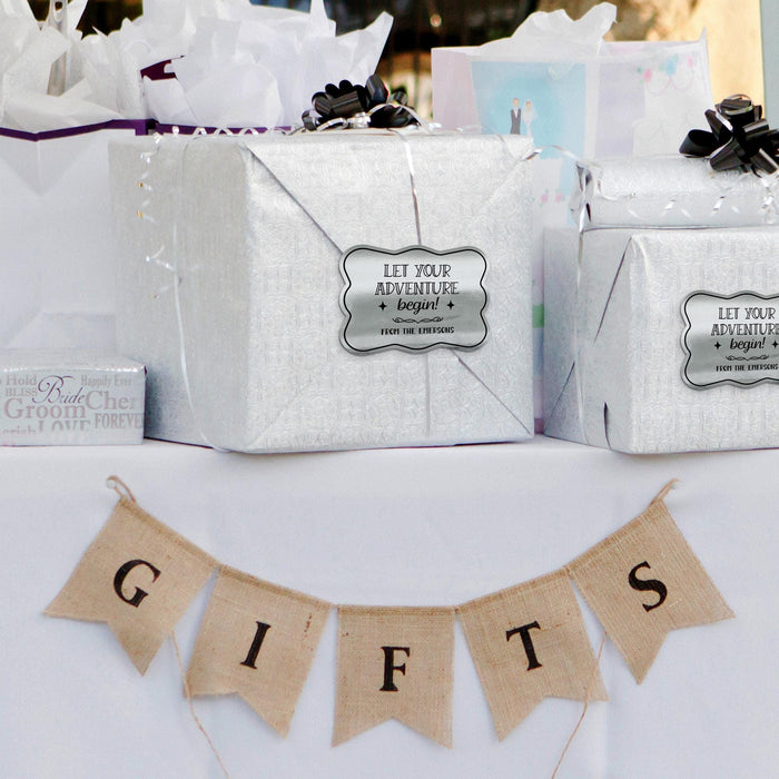 Silver gift bags decorated with personalized “Let Your Adventure Begin” metallic silver foil wedding gift tags. Styled with wedding décor and hanging banner accents for a romantic presentation.
