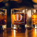 Warm bar-style lifestyle image with glowing ambient lights in the background. The personalized glass is filled with whiskey and ice, highlighting the elegant engraving.