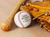 Baseball, wooden bat, and glove sits on wooden surface. Baseball has First Father's Day design engraved on it.