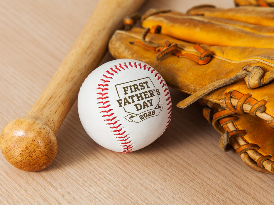 Baseball, wooden bat, and glove sits on wooden surface. Baseball has First Father's Day design engraved on it.