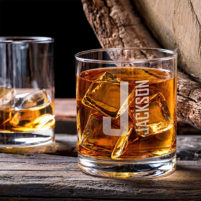 Lifestyle image of the whiskey glass with large ice cubes on a wooden table beside another drink. The custom engraving adds a stylish and personalized bar feel.