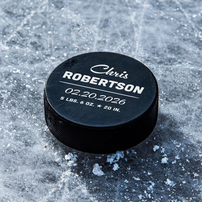 Personalized black hockey puck with a white UV printed birth announcement design featuring a script first name, bold last name, birth date, weight, and length.