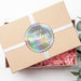 Personalized round Happy Holidays gift tag displayed on a kraft gift box with white ribbon and pink crinkle paper.