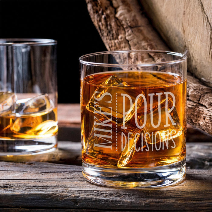 Lifestyle image of the personalized whiskey glass with whiskey and large ice cubes placed on a wooden table beside another drink. The custom engraving adds a refined and personalized bar-inspired look.