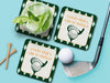 Custom square pulpboard coasters are shown sitting on a blue surface. Golf tees, a golf ball, and a golf club sit beside them. One coaster has a cocktail on top of it. The coaster features design with an illustration of a golf ball and with club behind it, argyle background pattern, and custom text.