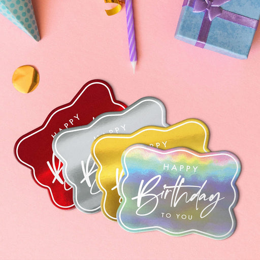 Four elegant script birthday tags in metallic gold, metallic silver, metallic red, and metallic rainbow lying beside colorful gifts and party decorations.