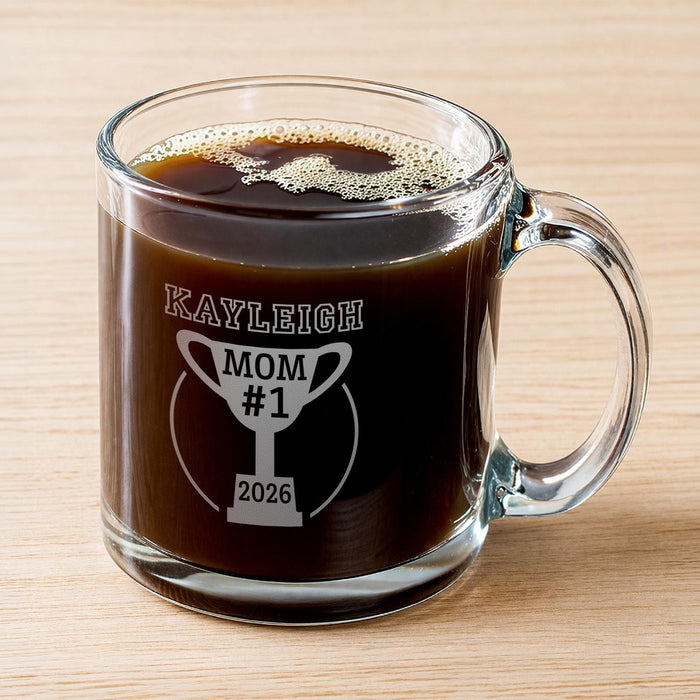 A glass mug filled with a dark liquid, likely coffee, with foam on top. The mug has a personalized message printed on it, which reads "Kayleigh Mom #1 2026".