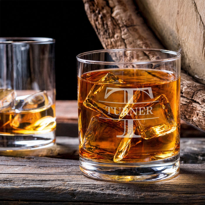 Lifestyle image of the whiskey glass with large ice cubes on a wooden table beside another drink. The custom engraving adds a refined and personalized bar feel.