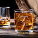 Lifestyle image of the whiskey glass with large ice cubes on a wooden table beside another drink. The custom engraving adds a refined and personalized bar feel.