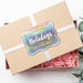 Personalized rounded rectangle Happy Holidays gift tag displayed on a kraft gift box with white ribbon and pink crinkle paper.