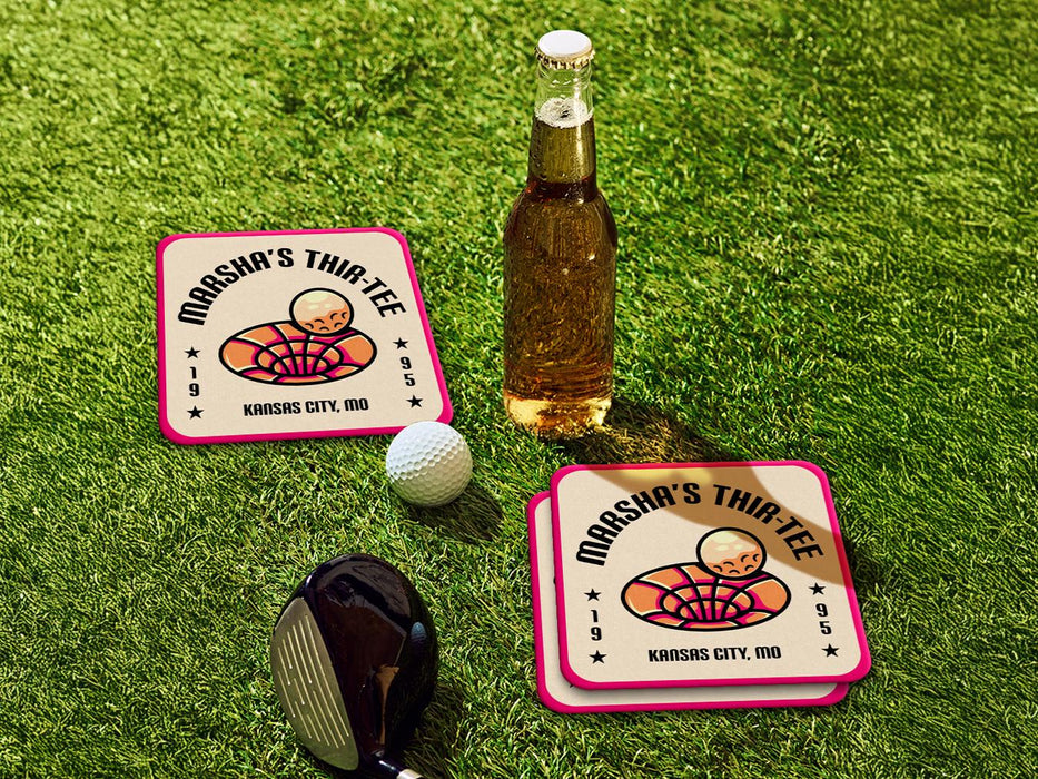 Custom square pulpboard coasters are shown sitting on a grassy, green golf course next to a bottle of beer, white golf ball, and golf club. The coaster features design with illustrated golf ball and hole, custom name, age, year, and location, and vivid orange, cream, and pink color palette.