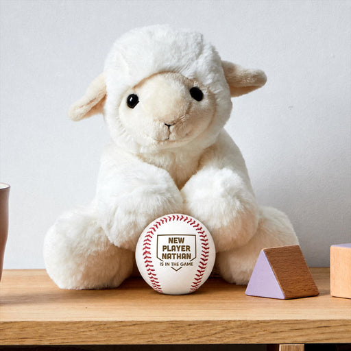 Personalized engraved New Player baseball birth announcement displayed beside a plush lamb and baby nursery decor on a wooden shelf.