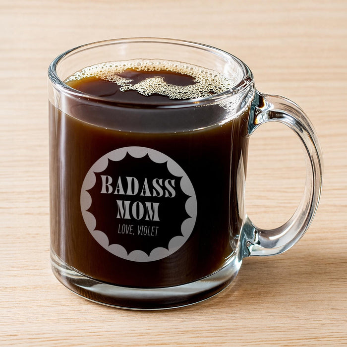 A glass mug filled with a dark liquid, likely coffee, with foam on top. The mug has an engraved design on it that says "BADASS MOM" and "LOVE. VIOLET".