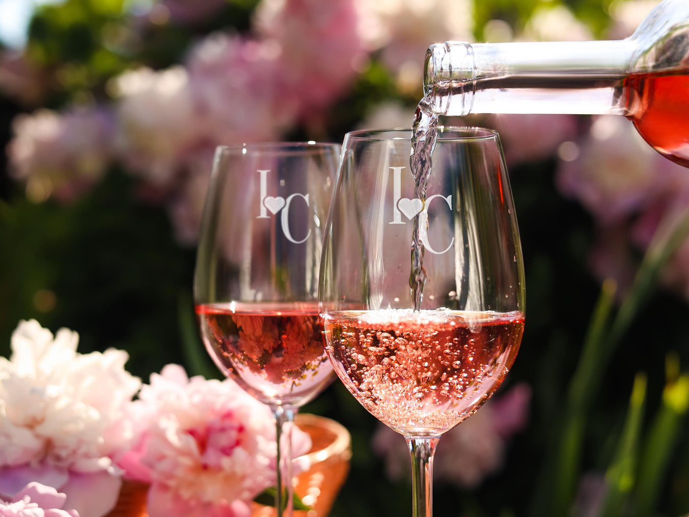 Two wine glasses sit in front of flowers as wine is being poured into them. The glasses have Heart Initial design engraved on them.