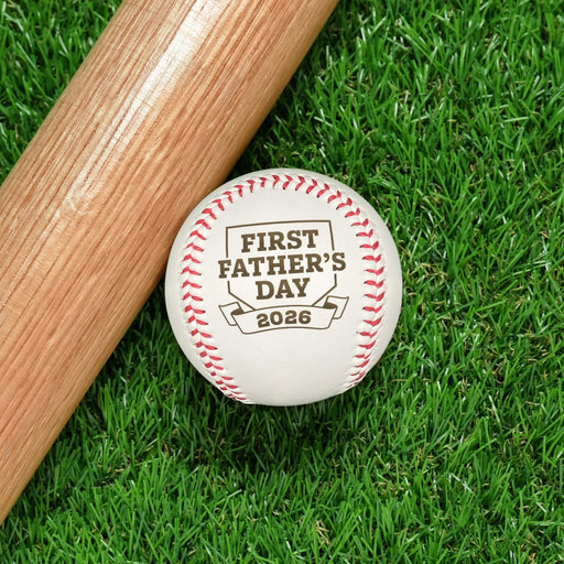 Baseball sits on grass next to baseball bat. Baseball has First Father's Day design engraved on it.