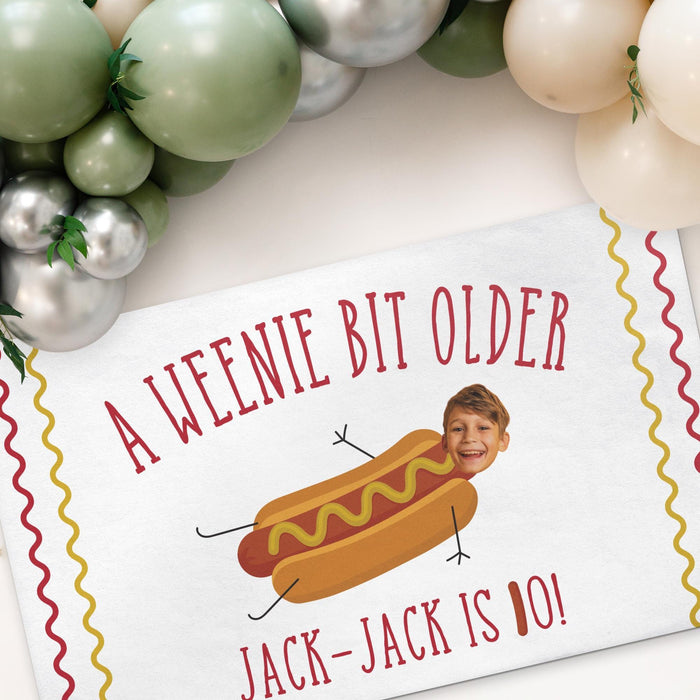 Personalized hot dog birthday banner displayed beneath a balloon garland featuring a custom name, age, and face cutout.