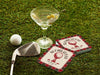 Custom square pulpboard coasters are shown sitting on a grassy, green golf course next to a martini glass, white golf ball, and golf club. The coasters feature design with an illustration of a golf ball on a tee, checkerboard border, and custom text.