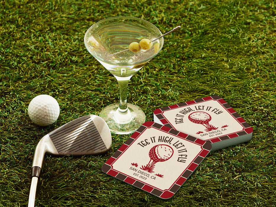 Custom square pulpboard coasters are shown sitting on a grassy, green golf course next to a martini glass, white golf ball, and golf club. The coasters feature design with an illustration of a golf ball on a tee, checkerboard border, and custom text.