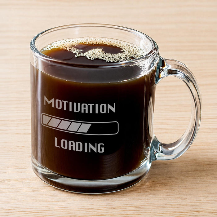 A glass mug filled with a dark liquid, likely coffee, with foam on top. The mug has an engraved message that reads "Motivation Loading".