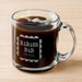 A glass mug filled with a dark liquid, likely coffee, with foam on top. The mug has an engraved message that says "BADASS DAD FROM RUSSEL".