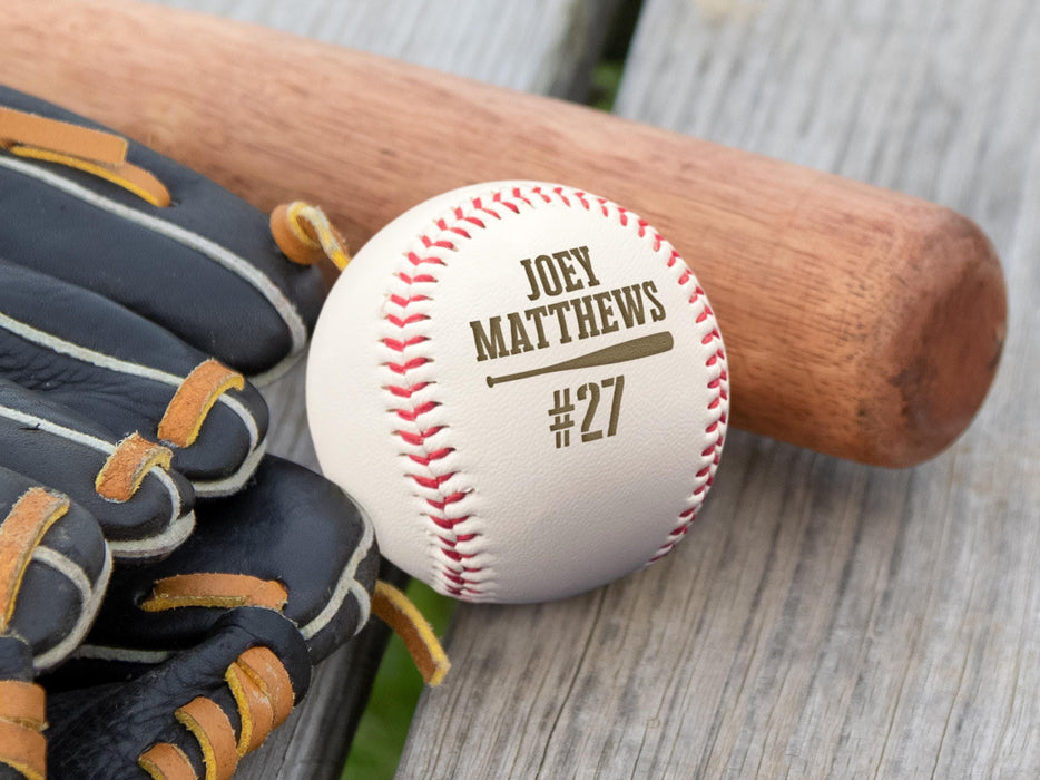 An engraved baseball, wooden bat, and baseball glove sit on a wooden surface outside. The engraved design features the player's name and jersey number along with a baseball bat graphic.