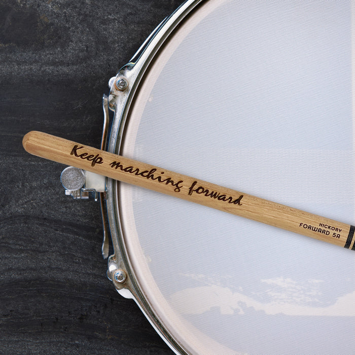 A drumstick with the words "Keep marching forward" engraved on it, resting on a drum set.