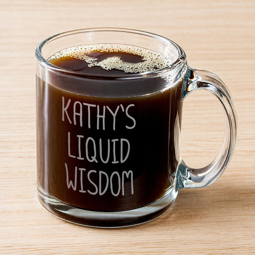 A glass mug filled with a dark liquid, likely coffee, with foam on top. IT has the engraving that says "Kathy's Liquid Wisdom". The mug has a handle and is placed on a wooden surface.
