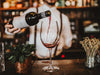 One wine glass sits on bar while bartender pours wine into it. The glass has Last Name and Established Date design engraved on it.