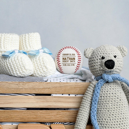 Personalized engraved baseball birth announcement shown with blue baby booties, a knitted teddy bear, and nursery decor.