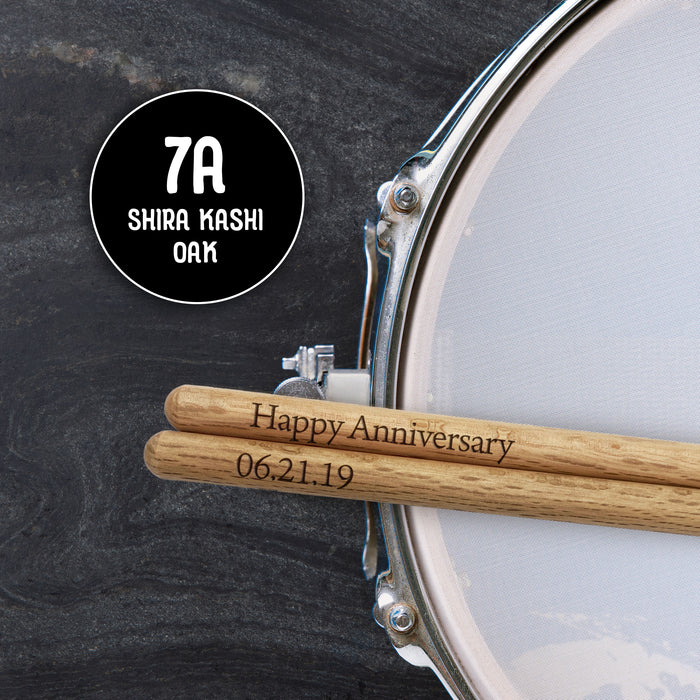 A pair of drumsticks with the text "Happy Anniversary" engraved on them, placed on a drum set.