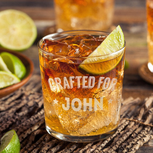 Personalized whiskey glass displayed with whiskey, ice, and fresh lime on a rustic wooden surface. The “Crafted For John” engraving is clearly visible, showing how the finished custom rocks glass looks in a stylish home bar setting.