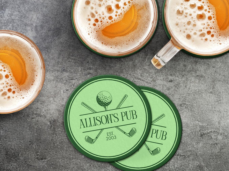 Custom round pulpboard coasters are shown sitting on a dark gray stone background with beer mugs surrounding them. The beer mugs have coasters underneath them. The custom coasters feature a green, elegant golf bar design with interlocking golf clubs and a golf ball sitting on top of a golf tee. The bar name and established year can be customized.