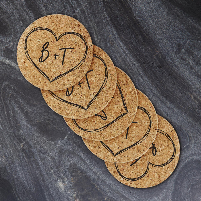 Set of personalized cork coasters with large hand-drawn heart monograms arranged on a dark fabric background.