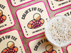 Custom square pulpboard coasters are shown laid out on a wooden surface. A beer mug is shown on top of a few of the coasters. The coaster features design with illustrated golf ball and hole, custom name, age, year, and location, and vivid orange, cream, and pink color palette.