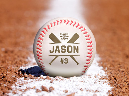 An engraved baseball sits on an orange clay baseball field. This engraved design features a bold crossed bat graphic with a baseball player's graduating year, first name, and jersey number.