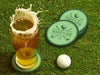 Custom round pulpboard coasters are shown sitting on a grassy, green golf course next to a pint of beer and a white golf ball. The pint of beer has a golf ball splashing into it and has a coaster underneath it. The custom coasters feature a green, elegant golf bar design with interlocking golf clubs and a golf ball sitting on top of a golf tee. The bar name and established year can be customized.