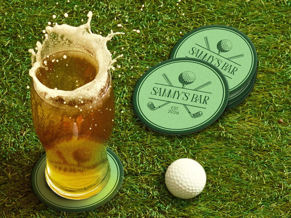 Custom round pulpboard coasters are shown sitting on a grassy, green golf course next to a pint of beer and a white golf ball. The pint of beer has a golf ball splashing into it and has a coaster underneath it. The custom coasters feature a green, elegant golf bar design with interlocking golf clubs and a golf ball sitting on top of a golf tee. The bar name and established year can be customized.