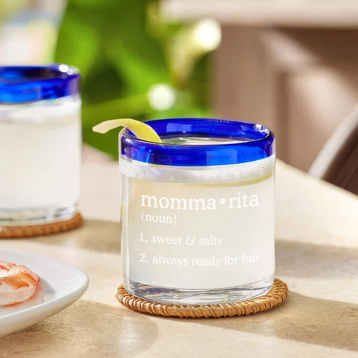 A glass with a blue rim and a lemon twist. The engraving says "momma rita noun" & "1 sweet & salty 2 always ready for fun". This glass sits on a table with a plate of food in the background.