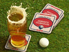Custom square pulpboard coasters are shown sitting on a grassy, green golf course next to a pint of beer and a white golf ball. The pint of beer has a golf ball splashing into it and has a coaster underneath it. The custom coasters feature a 19th hole bar design with an illustration of a golf ball. The tagline, bar name, established year, and location can be customized on these.