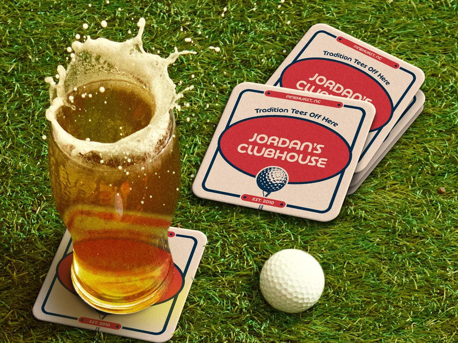 Custom square pulpboard coasters are shown sitting on a grassy, green golf course next to a pint of beer and a white golf ball. The pint of beer has a golf ball splashing into it and has a coaster underneath it. The custom coasters feature a 19th hole bar design with an illustration of a golf ball. The tagline, bar name, established year, and location can be customized on these.