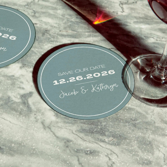 Personalized Save the Date coasters displayed beside a wine glass. Features a modern circular design with custom names and wedding date.