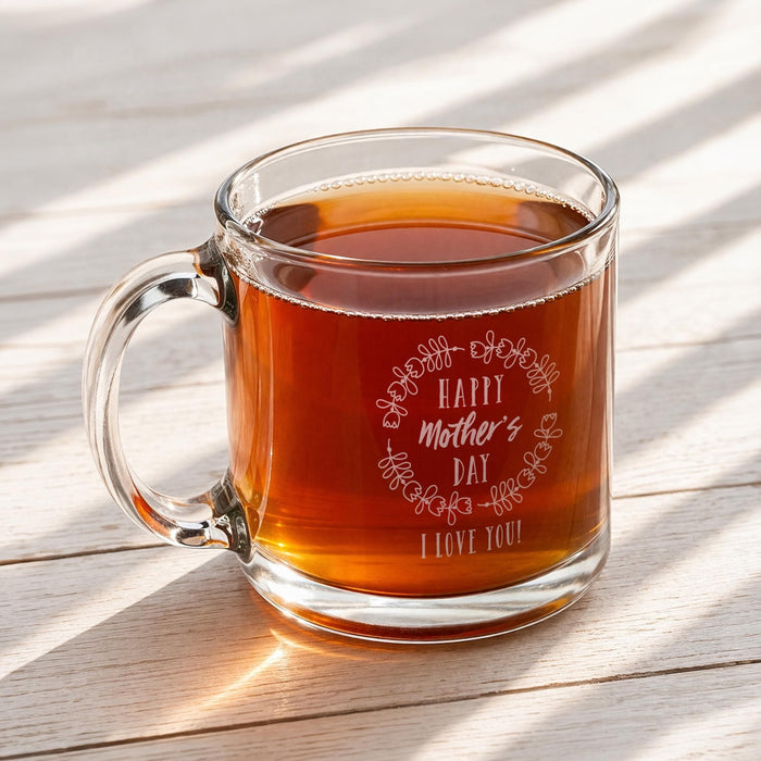 A glass mug filled with a warm, amber-colored liquid, likely tea, sits on a wooden surface. The mug has a message engraved on it that reads "Happy Mother's Day I love you!"